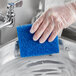 A hand in clear plastic gloves using a blue Lavex multi-purpose scouring pad to clean a sink.