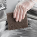 A hand in a glove using a brown Lavex multi-purpose scouring pad to clean a sink.