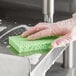 A hand in a clear plastic glove cleaning a Lavex green cellulose sponge on a stainless steel sink.