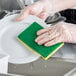 A hand in a glove using a green Lavex sponge to clean a plate.