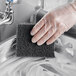 A hand in a clear plastic glove scrubbing a sink with a black Lavex multi-purpose scouring pad.