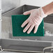 A hand in a plastic glove using a Lavex medium-duty dark green scouring pad to clean a dishwasher.
