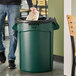 A person standing next to a Rubbermaid green BRUTE trash can.