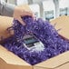 A hand holding a package in a cardboard box with purple shredded paper.