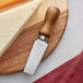 An Acopa stainless steel cheese knife with a dark wood handle next to a piece of cheese on a cutting board.