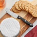 An Acopa stainless steel cheese spreader next to crackers on a cutting board.