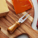 An Acopa stainless steel cheese fork with a dark wood handle on a cutting board with cheese.