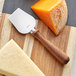 An Acopa stainless steel cheese knife with a natural pakkawood handle on a cutting board with a piece of cheese.