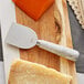 An Acopa stainless steel cheese knife with a white marble handle cutting a block of cheese.