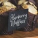 A table with blueberry muffins in brown wrappers with a white mini chalk card sign that says "blueberry muffins"