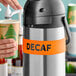A hand using a white silicone label band to label a stainless steel airpot as "Decaf"