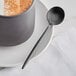 An Acopa Odin stainless steel demitasse spoon on a plate with a cup of coffee.