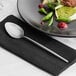 An Acopa Odin stainless steel dinner spoon on a black napkin next to a plate of food.