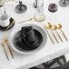 A table setting with a gold napkin folded on a black plate and an Acopa Odin gold dinner knife and spoons.
