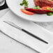 An Acopa Odin stainless steel dinner knife on a napkin next to a plate of meat and vegetables.