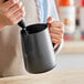 A hand using a black Acopa frothing pitcher to pour milk.