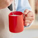 A person using a red Acopa frothing pitcher to pour milk into a mug.