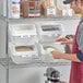 A man in a red apron uses a scoop to fill a Baker's Mark ingredient shelf bin with rice.