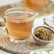 A glass cup of Davidson's Organic Green Tea Garden tea with a strainer of leaves and flowers.