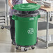 A woman pushing a Lavex green round commercial recycling can with a green lid and dolly.