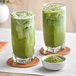 Two glasses of David Rio Super Blends Matcha Latte with a bowl of green powder.
