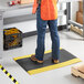 A person wearing blue jeans standing on a Lavex Diamond Deluxe black anti-fatigue mat with yellow borders.