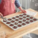 A woman wearing a red apron uses a Choice aluminum muffin and cupcake pan to bake brown cupcakes.