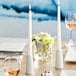 A table set with a white tablecloth and a white Acopa taper candle holder with a lit candle.
