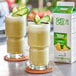 Two glasses of green smoothie with cucumber slices and a carton of Island Oasis Plus Tropical Greens Frozen Beverage Mix.