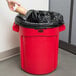 A hand throwing a coffee cup into a Rubbermaid red trash can with a black bag.