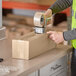 A person wearing a safety vest and using a tape dispenser to seal a Lavex Kraft corrugated shipping box.