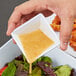A hand holding a square white bowl with yellow sauce being poured over a salad.