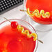 A pair of glasses with red and yellow cocktails, each garnished with a Barfly gold cocktail pick with a spiral on it.