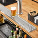 A Regency stainless steel surface mount beer drip tray on a bar counter with a glass of beer and liquor bottles.