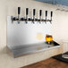 A Regency stainless steel wall mount beer drip tray with six faucets over a glass of beer.