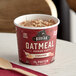 A red Kodiak Cakes Cinnamon Oatmeal cup on a table with a bowl of oatmeal and a wooden spoon.