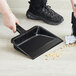 A person using a black Lavex plastic utility dust pan with a handle to sweep the floor.