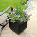 A Mayne black planter with purple flowers and green grass on an outdoor patio table.