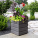 A Mayne graphite gray planter with flowers in it on a sidewalk.