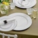 A table set with white Choice charger plates and silverware with a white napkin on a plate.