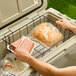 A person is placing a wire basket containing packaged hot dog buns and hot dogs into a large cooler with ice and beverages inside.