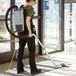 A woman wearing a ProTeam backpack vacuum with a wand and floor tool vacuums a grocery store floor.
