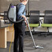 A woman with a ProTeam backpack vacuum cleaning the floor of a corporate office with green chairs.