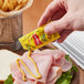 A hand holding a French's classic yellow mustard packet over a ham sandwich on a table in a deli.