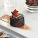 A chocolate cake on a white plate with a piece of Valrhona dark chocolate on top.