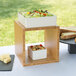 A white square container of pasta and a white box with salad on a bamboo cube riser on a table outdoors.