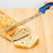 A Mercer Culinary Millennia Colors bread knife with a blue handle cutting a piece of bread on a cutting board.