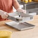 A person wearing gloves is holding a square metal frame above a rectangular metal baking sheet on a wooden countertop.