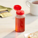 A 5.5 oz. clear PET sauce bottle with a red cap on a table next to a cup of tea.