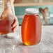 A person pouring honey into a classic queenline glass honey jar on a counter.